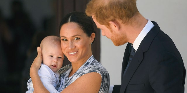 Meghan Markle in a printed dress holds Prince Archie as an infant as Prince Harry looks at them while in South Africa