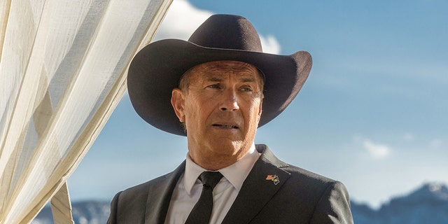Kevin Costner in a black suit and black tie looks off in the distance wearing a cowboy hat for 'Yellowstone' photo as John Dutton