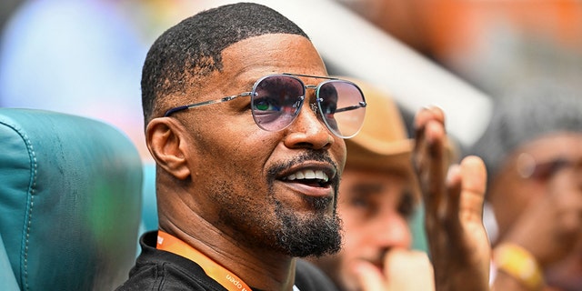Jamie Foxx raises his hand and waves with an orange lanyard around his neck wearing a black shirt while watching tennis in Miami