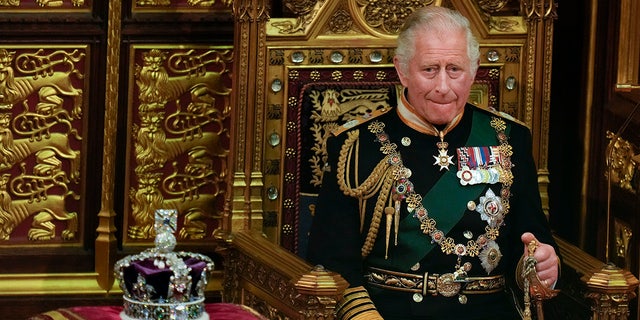 King Charles in green suit adorned with medals and chains sits in a royal chair next to a crown