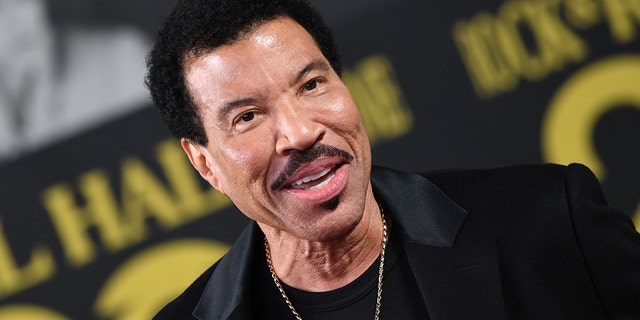 Lionel Richie smiles with a gold chain around his neck, velvet black suit and black shirt