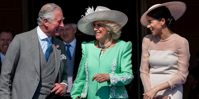 King Charles in a grey multi-piece suit smiles at Meghan Markle in a blush dress and hat, in the middle Camilla smiles in a bright green dress and white hat, looking at Charles