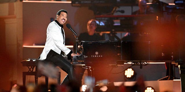Lionel Richie on stage playing the piano looks out to the crowd at the coronation concert, wearing a white jeweled jacket