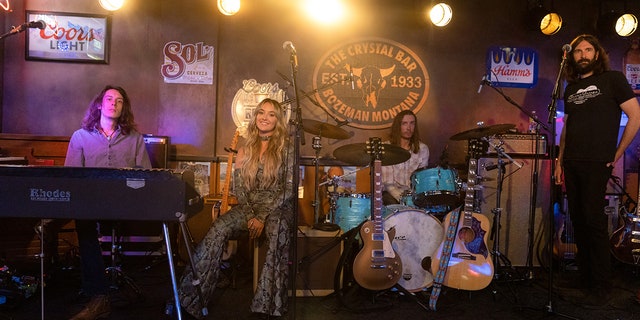 Lainey Wilson as Abby on "Yellowstone" in a black snakeskin jumpsuit sitting on stage smiling at the camera alongside her band mates