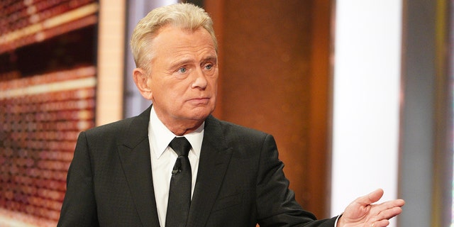 Pat Sajak in a black suit, white button down, and black tie looks off-camera while filming "Celebrity Wheel of Fortune"