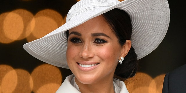 Meghan Markle smiles slightly off-camera in a white hat and matching outfit and earrings