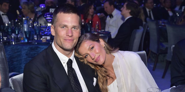 Gisele Bündchen in a white shirt and cream cardigan puts her head on Tom Brady's shoulder in a suit