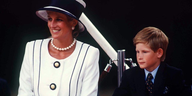 Princess Diana in a white suit with large buttons and a pearl necklace and matching top hat with a large black ribbon smiles nex to a young Prince Harry in a black suit and blue button down