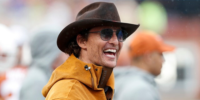Matthew McConaughey in an orange jacket laughs with a brown cowboy hat on and sunglasses