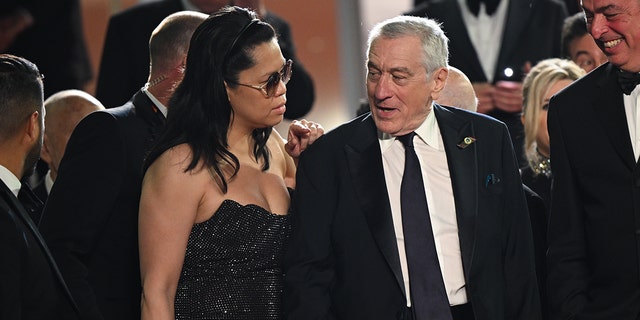 Tiffany Chen in a black sparkly gown and oversized sunglasses stands slightly behind Robert De Niro, who looks back to say something to her