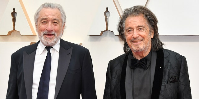 Robert De Niro smiles in a black suit and tie next to Al Pacino in a black suit at the Academy Awards in 2020