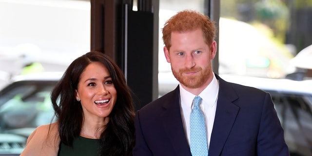 Meghan Markle in a green dress and camel colored jacket walks with Prince harry in a navy suit and teal tie