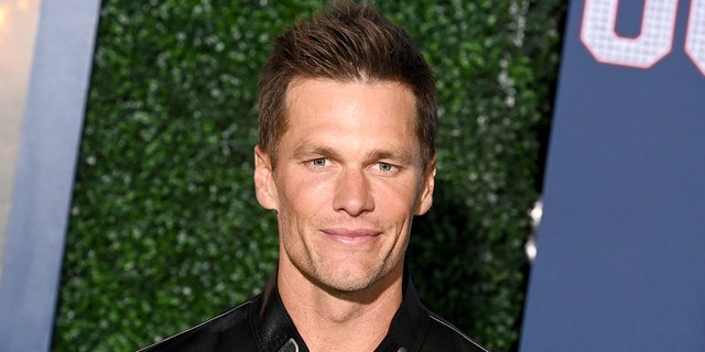 Tom Brady in a white shirt and leather jacket soft smiles and stares right at the camera on the "80 For Brady" red carpet