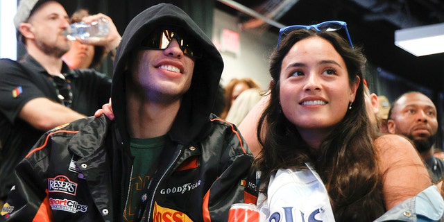 Pete Davidson in a black sweatshirt with his hood up and sunglasses on smiles next to Chase Sui Wonders at the Daytona 500 race in Florida