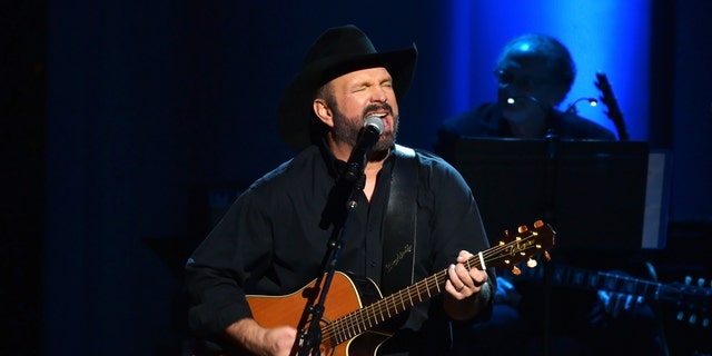 Garth Brooks performs on stage with a guitar in all black outfit