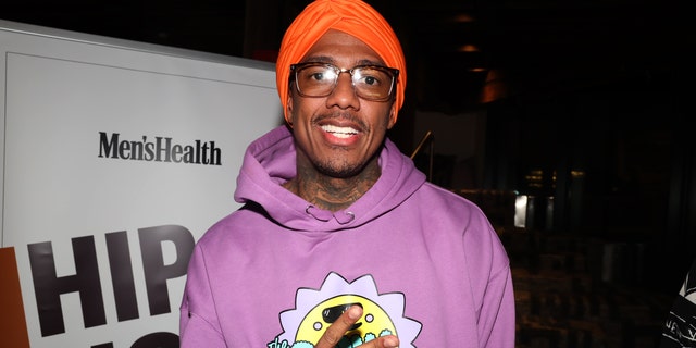 Nick Cannon in a purple sweatshirt throwing up a piece sign and wearing a red cap