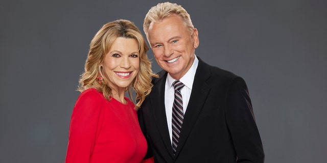 Vanna White in a red dress smiles in a promotional photo for "Celebrity Wheel of Fortune" with Pat Sajak in a black suit and striped tie