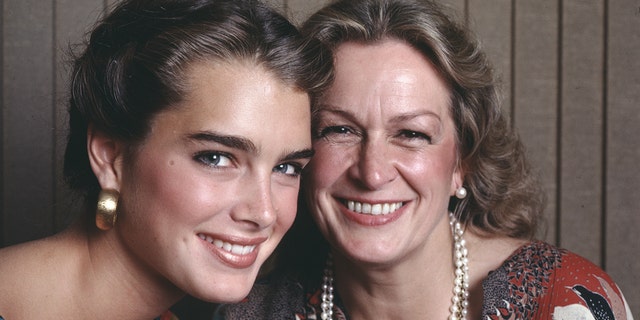 Brooke Shields in a colorful top and large gold earrings smiles next to her mother Teri Shields wearing a pearl necklace and earrings in a photo from 1981