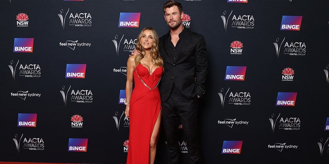 Chris Hemsworth in a black suit and jacket poses on the red carpet next to Elsa Pataky in a busty red gown in Sydney, Australia