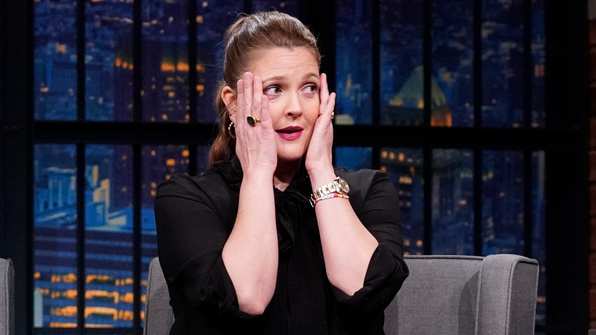 Drew Barrymore in a black dress puts her hands up to her face in distress on "Late Night with Seth Meyers"