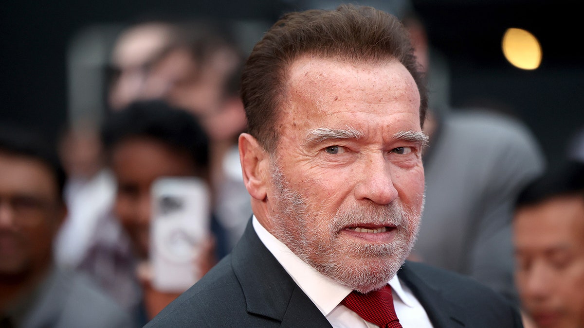 Arnold Schwarzenegger looks serious on the carpet in a dark suit and red tie