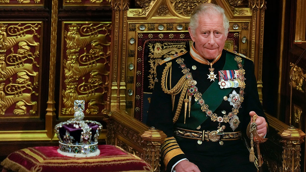 King Charles in green suit adorned with medals and chains sits in a royal chair next to a crown