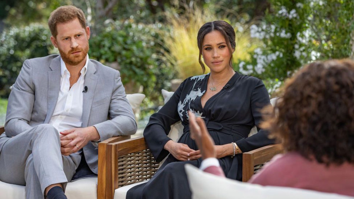 Prince Harry in a grey suit and white shirt sits next to wife Meghan Markle in a blue dress with light blue detail, across from the back of Oprah Winfrey doing an interview