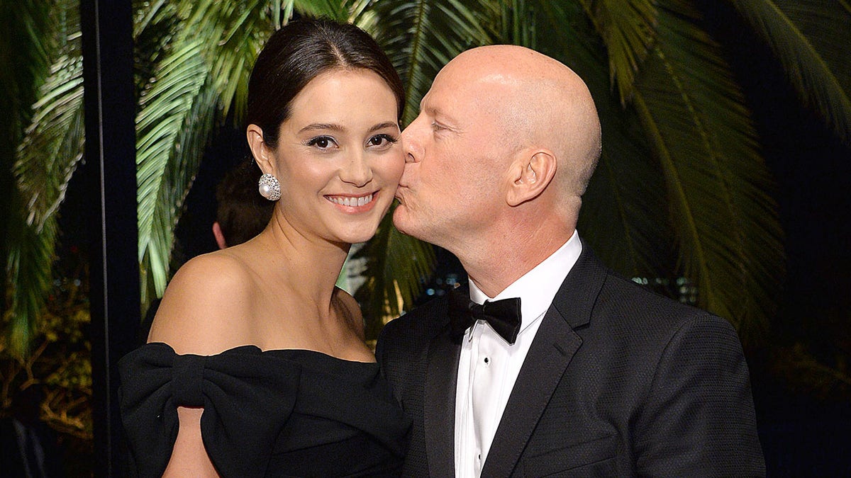 Bruce Willis in a classic tuxedo kisses wife Emma Heming Willis on the cheek, in a black dress