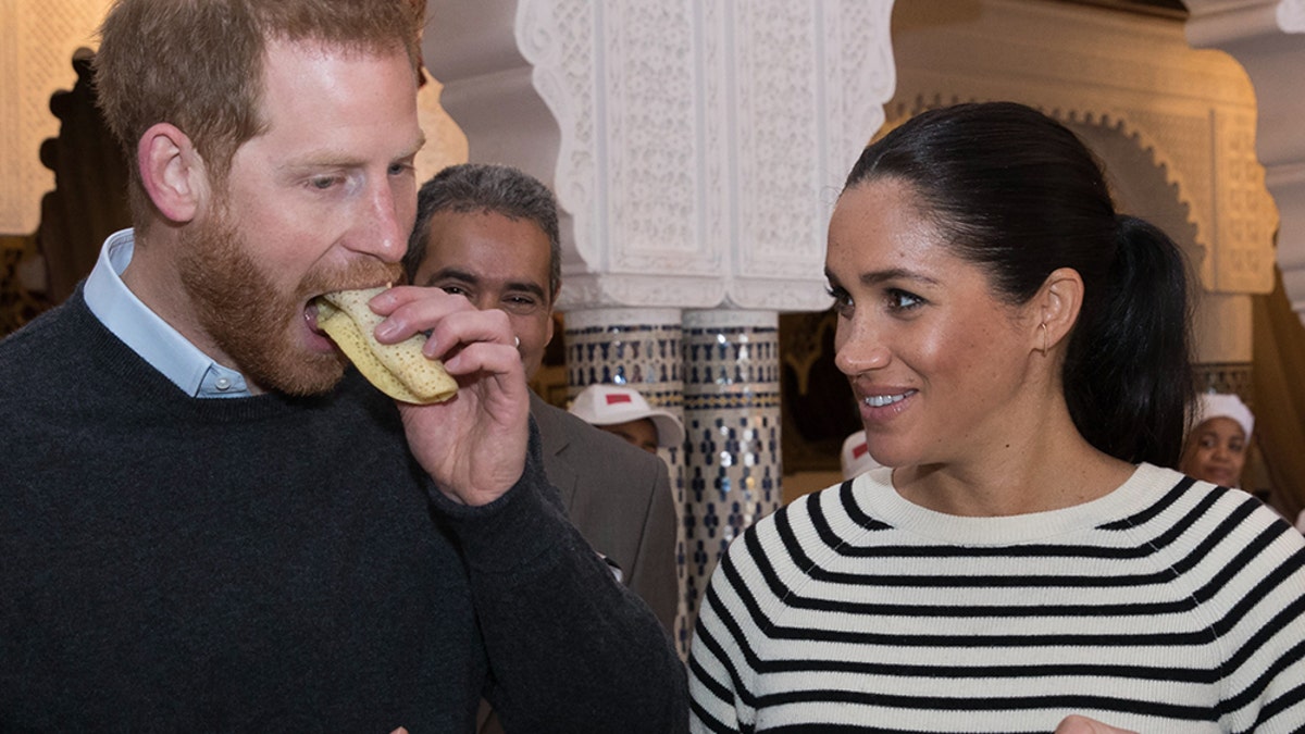 Prince Harry eating a sandwich as Meghan Markle looks on holding a plate