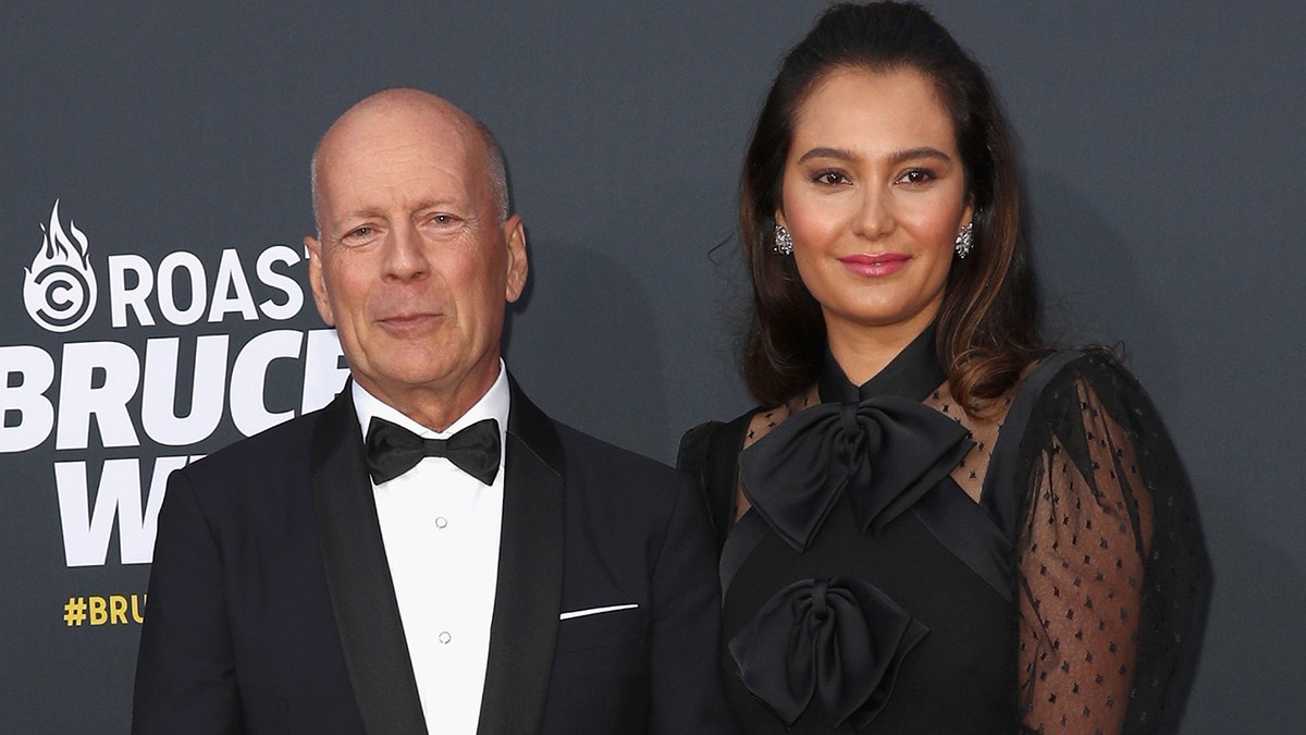 Bruce Willis in a classic tuxedo soft smiles for a picture with Emma Heming Willis on the carpet in Los Angeles
