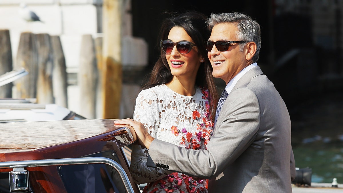 Amal Clooney and George Clooney wearing sunglasses smiling on a boat in Venice