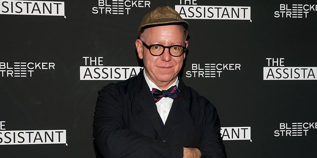 Writer James Schamus soft smiles for the camera at a screening of "The Assistant" wearing a suit and bow-tie, with his arms crossed, dark rimmed glasses and a hat