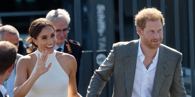 Meghan Markle waves in a white halter top and light pink pants while holding Prince Harry's hand in a grey suit