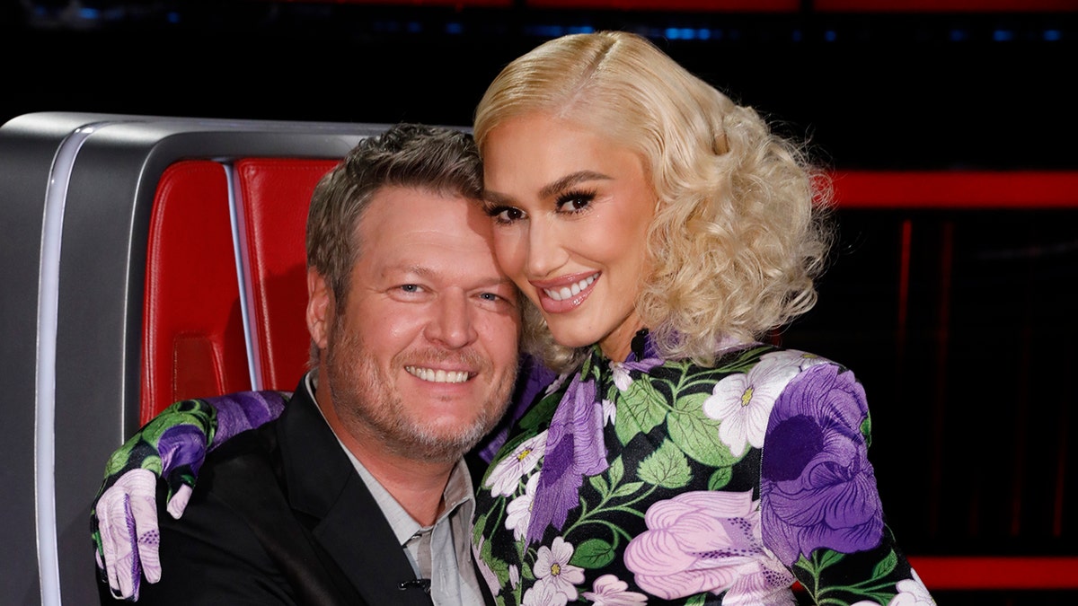 Blake Shelton in a black suit jacket and shirt hugs wife Gwen Stefani in a purple, green, and white floral outfit