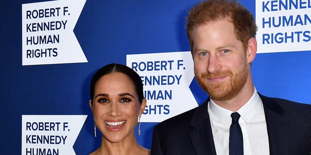 Meghan Markle smiles in a white dress and dangly earrings next to Prince Harry who soft smiles in a navy suit at the Robert F. Kennedy Human Rights Ripple of Hope Award Gala red carpet