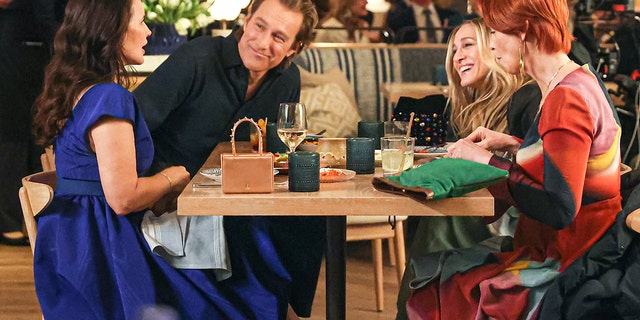 Kristin Davis (Charlotte) John Corbett (Aidan) Sarah Jessica Parker (Carrie) and Cynthia Nixon (Miranda) sit at a table and laugh while filming a scene for "And Just Like That"