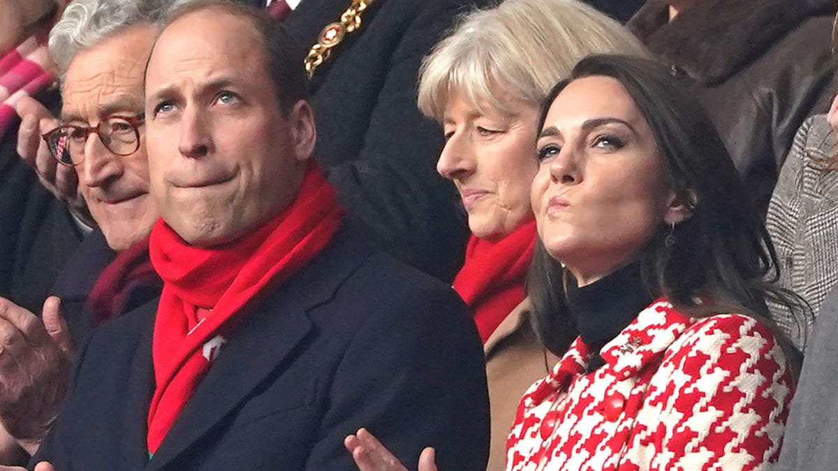 Prince William in a dark long jacket and bright red solid scarf in the stands alongside his wife Kate Middleton in a patterned red and white jacket at the Wales vs. England rugby match