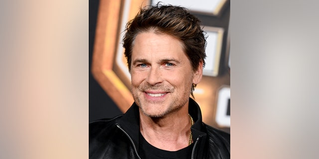 Rob Lowe smiles directly at the camera in a black jacket on the red carpet of "Guardians of the Galaxy Vol. 3"
