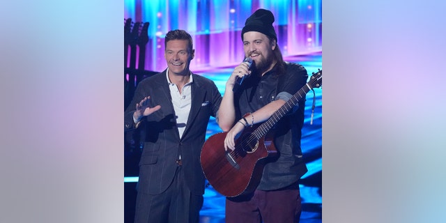 Host Ryan Seacrest stands on stage with "American Idol" contestant Oliver Steele in a beanie and holding a guitar