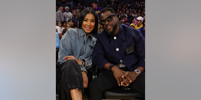 Eniko Hart in a denim jacket sits next to husband Kevin Hart wearing black sunglasses inside at the Lakers game
