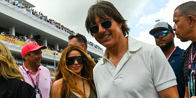 Shakira in a black tank top with two strings in the center around her neck and massive glasses soft smiles for a picture with Tom Cruise in a white polo shirt at the Grand Prix