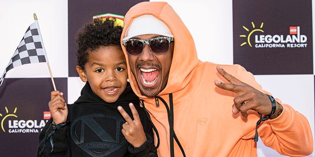 Nick Cannon in an orange sweatshirt, white band around his head and dark sunglasses poses with his son Golden Cannon doing a peace sign while wearing black on the red carpet