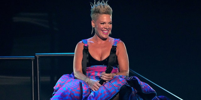 Pink sits on stage in a black top/printed bottom dress and smiles at the iHeartRadio Music Awards