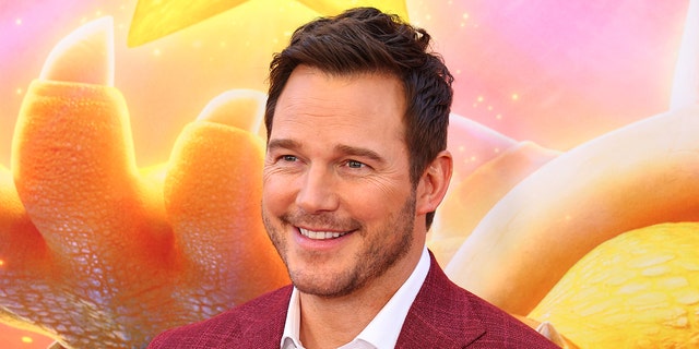 Chris Pratt wearing a white button down and red jacket smiles on the red carpet