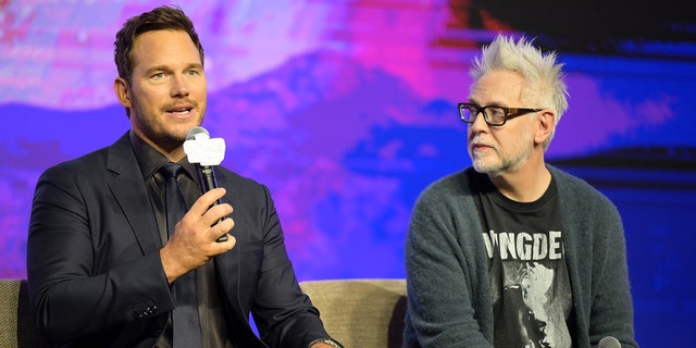 Chris Pratt wearing a blue suit and black shirt speaks into the microphone, sitting next to director James Gunn who is wearing a black t-shirt and blue sweater on stage