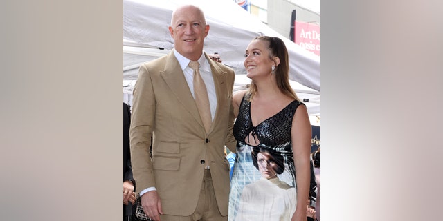 Bryan Lourd in a beige suit and white shirt and yellow/beige tie smiles as his daughter Billie Lourd looks lovingly up at him, with her hair in two pigtails, wearing a dress with her mother Carrie Fisher's face on it as Princess Leia from "Star Wars"