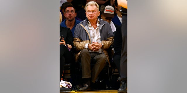 Pat Sajak sits courtside in a two-toned blue and tan raincoat with his hands clasped together
