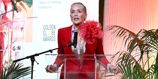 Sharon Stone holds the glass podium as she speaks to the "Raising Our Voices" luncheon crowd wearing a red suit with a flower