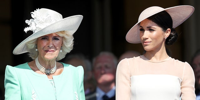 Queen Camilla wearing a green dress with a white hat standing next to Meghan Markle wearing a pink dress with a matching hat