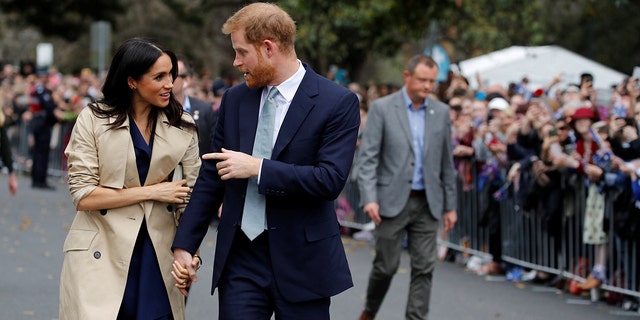 Meghan Markle wearing a trench coat and a black dress with prince harry wearing a dark suit and a light blue tie